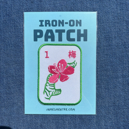 An iron-on patch displayed on a blue denim fabric background. The patch is on a light blue backing card with the text 'IRON-ON PATCH'. The patch features an embroidered design of a mahjong tile of a pink cherry blossom flower with green leaves on a white background, labeled with the number 1 and the Chinese character for plum.