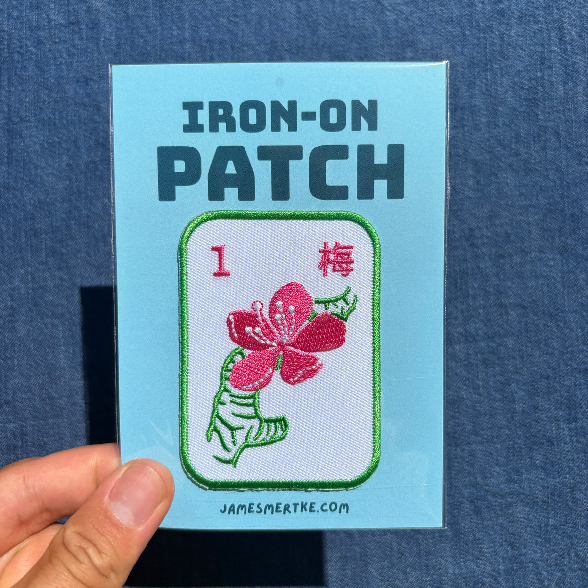 A hand holds a light blue backing card for an iron-on patch. The card reads 'IRON-ON PATCH' and shows a white embroidered patch with a pink flower, green leaves, a green border, and the number 1 with Chinese characters.