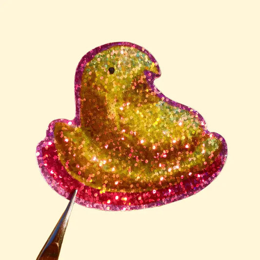 This sticker depicts a yellow marshmallow peeps treat with a pink outline. The sticker is made with a special material to give it a glittery and sparkly effect.