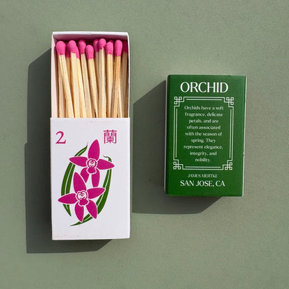 A matchbox is illustrated with a green and pink orchid to look like a mahjong tile. There is info about the meaning of the plant on the back. Matches with pink heads are inside the box.