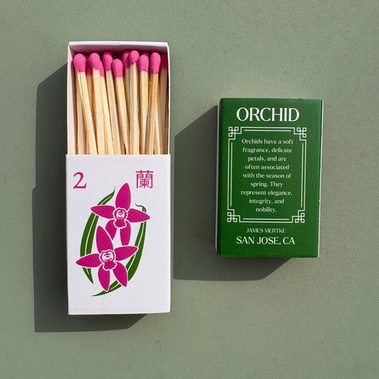 A matchbox is illustrated with a green and pink orchid to look like a mahjong tile. There is info about the meaning of the plant on the back. Matches with pink heads are inside the box.