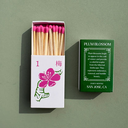 A matchbox is illustrated with a green and pink plum blossom to look like a mahjong tile. There is info about the meaning of the plant on the back. Matches with pink heads are inside the box.