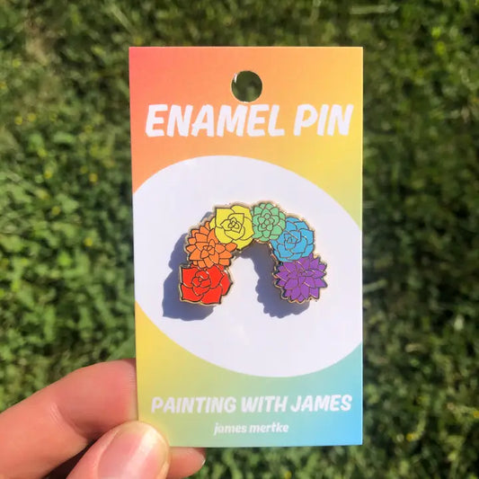 An enamel pin depicts 6 flowers or succulents in the shape of an arch. Each succulent is a different color of the rainbow.