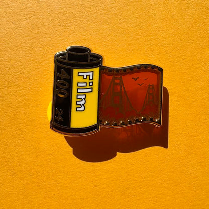 This pin features a film roll canister with "Film 400 24" written on it. It features a "stained glass" transparent panel with a screen-printed detail of San Francisco and the Golden Gate bridge when held up to the light.