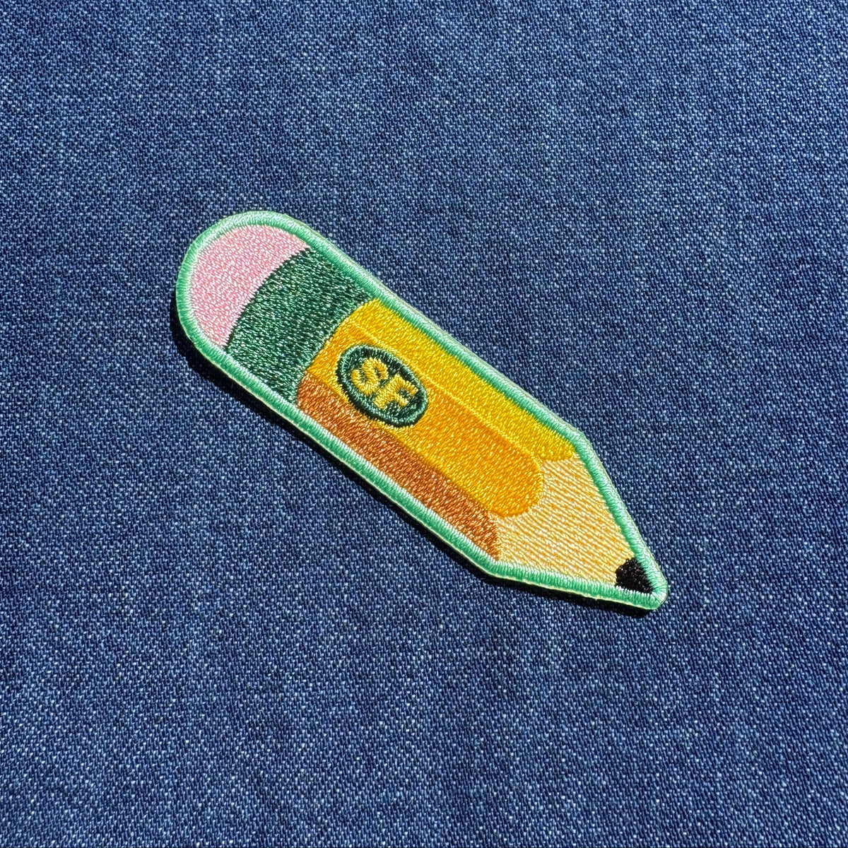 A colorful embroidered pencil-shaped patch with pink, green, yellow, and orange sections, placed on a blue denim fabric background.