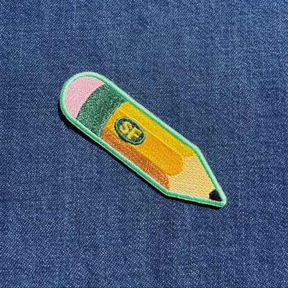 A colorful embroidered pencil-shaped patch with pink, green, yellow, and orange sections, placed on a blue denim fabric background.