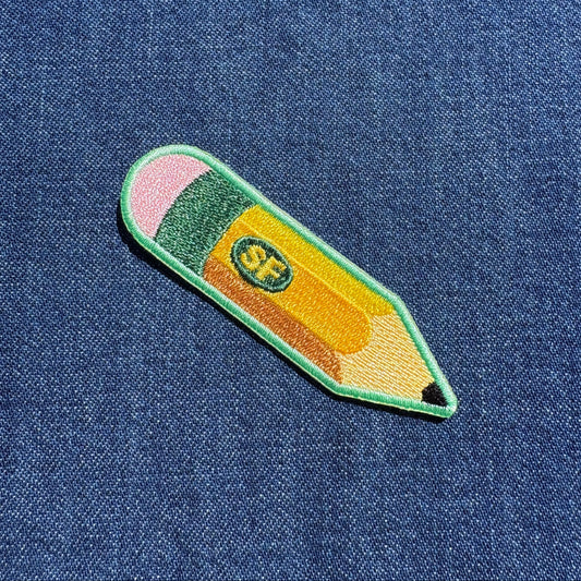 A colorful embroidered pencil-shaped patch with pink, green, yellow, and orange sections, placed on a blue denim fabric background.