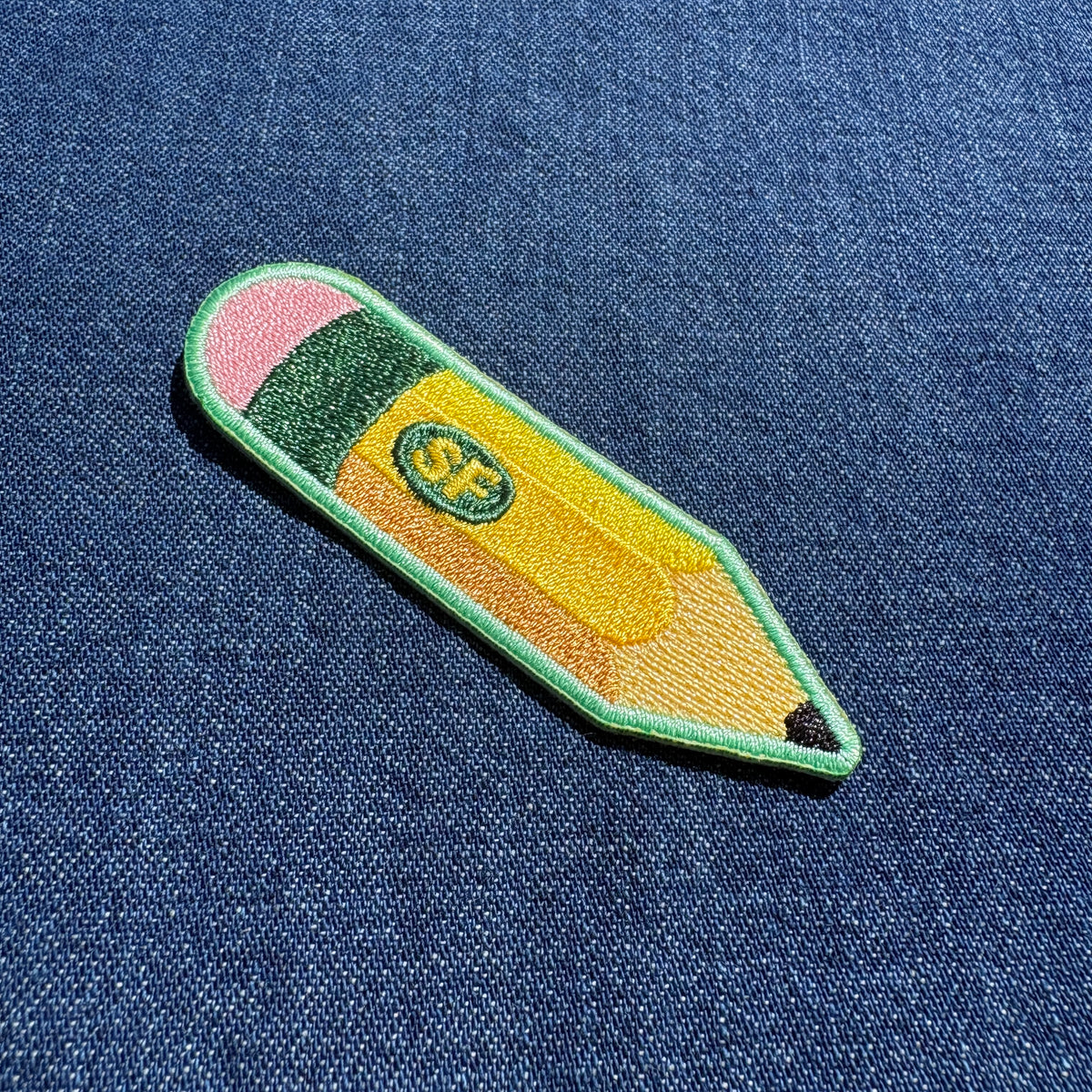 An embroidered patch shaped like a pencil with a green border, pink eraser, yellow body, and a small green oval containing the letters 'SF', placed on a blue denim background.