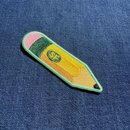 An embroidered patch shaped like a pencil with a green border, pink eraser, yellow body, and a small green oval containing the letters 'SF', placed on a blue denim background.