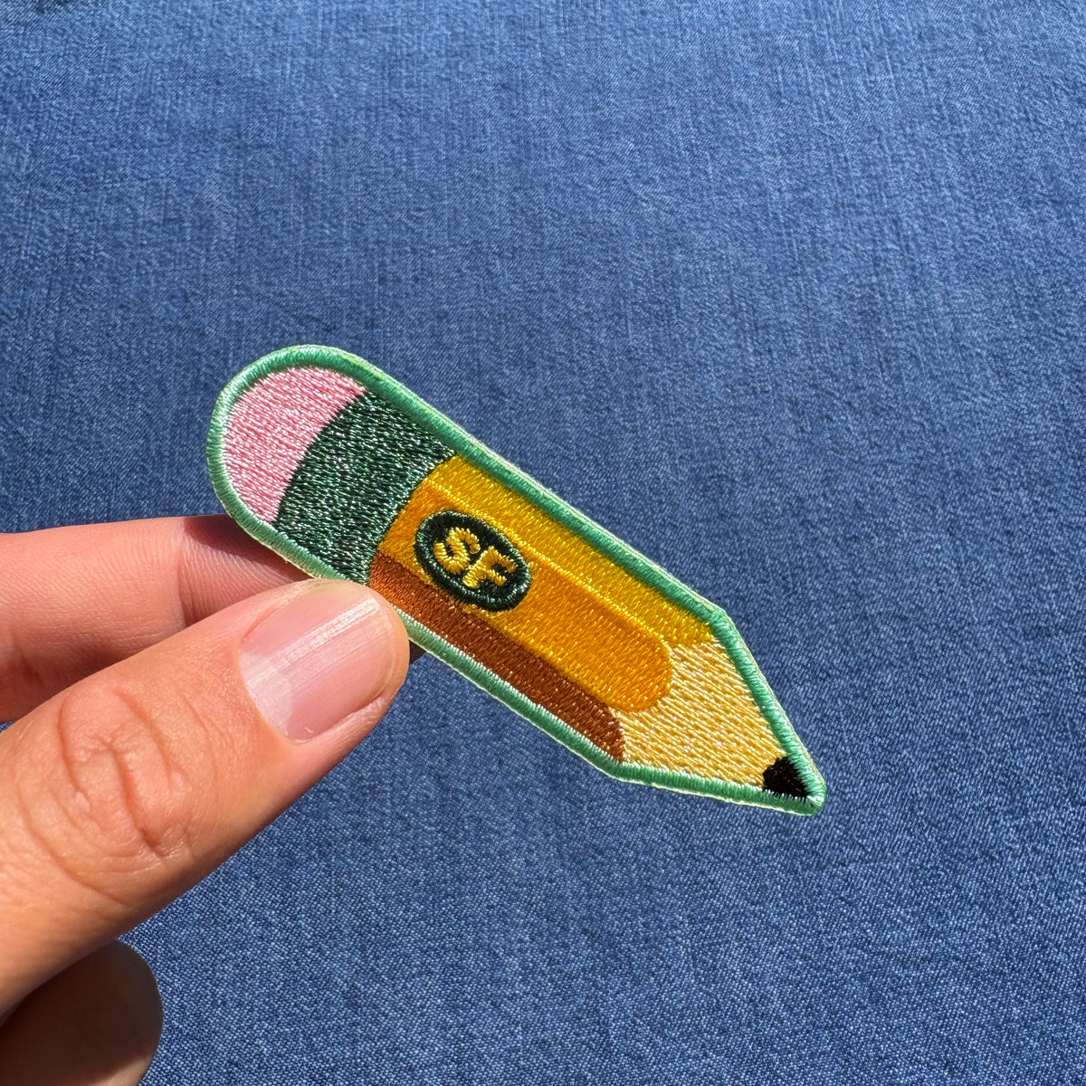 A hand holding an embroidered pencil patch against a blue textured fabric background. The patch is shaped like a yellow pencil with a green border, pink eraser, dark green middle section, and a small center logo reading 'SF'.