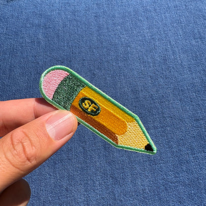 A hand holding an embroidered pencil patch against a blue textured fabric background. The patch is shaped like a yellow pencil with a green border, pink eraser, dark green middle section, and a small center logo reading 'SF'.