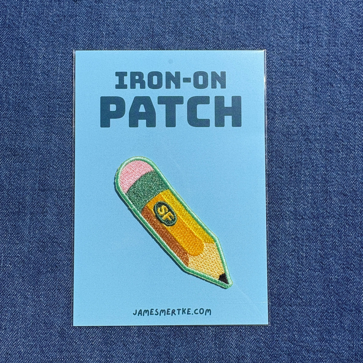 An embroidered iron-on patch shaped like a yellow pencil with a pink eraser and green tip, displayed on a light blue backing card that reads 'IRON-ON PATCH' and 'JAMESMERTKE.COM', set against a blue denim background.