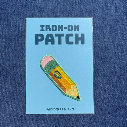 An embroidered iron-on patch shaped like a yellow pencil with a pink eraser and green tip, displayed on a light blue backing card that reads 'IRON-ON PATCH' and 'JAMESMERTKE.COM', set against a blue denim background.