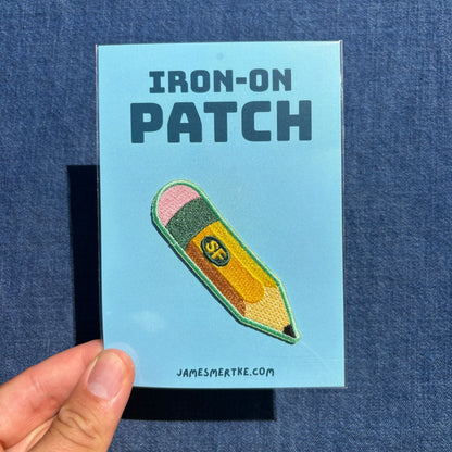 A hand holds a light blue backing card displaying an embroidered iron-on patch shaped like a yellow pencil with pink, green, and orange details. The card reads 'IRON-ON PATCH' and 'JAMESMERTKE.COM'