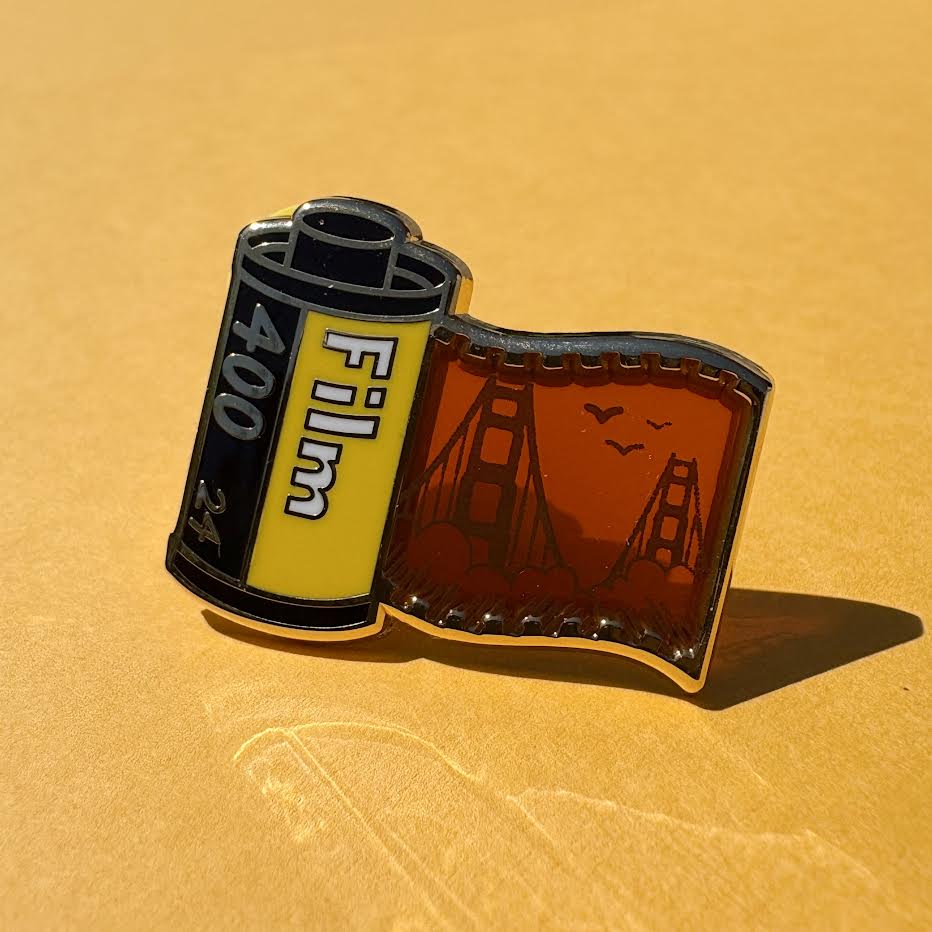 This pin features a film roll canister with "Film 400 24" written on it. It features a "stained glass" transparent panel with a screen-printed detail of San Francisco and the Golden Gate bridge when held up to the light.