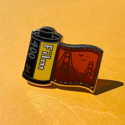 This pin features a film roll canister with "Film 400 24" written on it. It features a "stained glass" transparent panel with a screen-printed detail of San Francisco and the Golden Gate bridge when held up to the light.