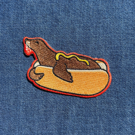 An embroidered patch of a sea lion in a hot dog bun with red outline, and yellow mustard placed on a blue denim fabric background.
