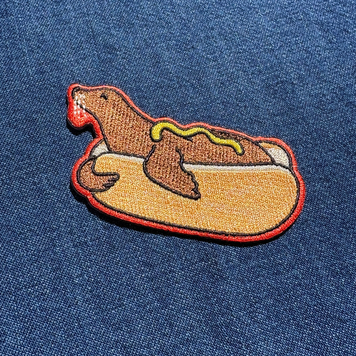 An embroidered patch of a sea lion dressed like a hot dog, featuring a brown seal in a tan bun with red, yellow, and white accents, shown on a blue denim fabric background.