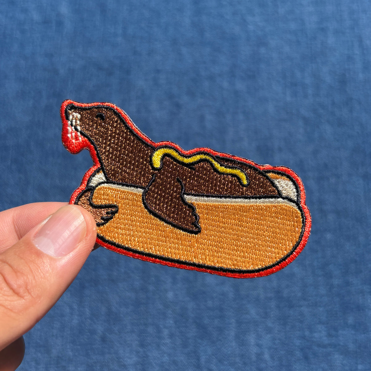 An embroidered patch shaped like a brown sea lion dressed like a hot dog, held by a person's fingers against a textured blue background.