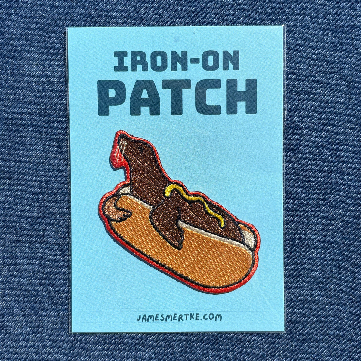 An iron-on patch featuring a brown sea lion dressed like a hot dog with mustard and ketchup, displayed on a light blue backing card that reads 'IRON-ON PATCH' and 'JAMESMERTKE.COM', laid flat on a blue denim background.
