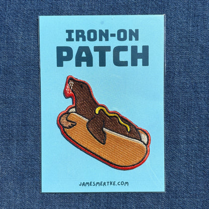 An iron-on patch featuring a brown sea lion dressed like a hot dog with mustard and ketchup, displayed on a light blue backing card that reads 'IRON-ON PATCH' and 'JAMESMERTKE.COM', laid flat on a blue denim background.