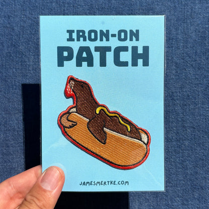 A hand holds a light blue card labeled 'IRON-ON PATCH' featuring an embroidered sea lion hot dog patch. The card also shows the website JAMESMERTKE.COM . 