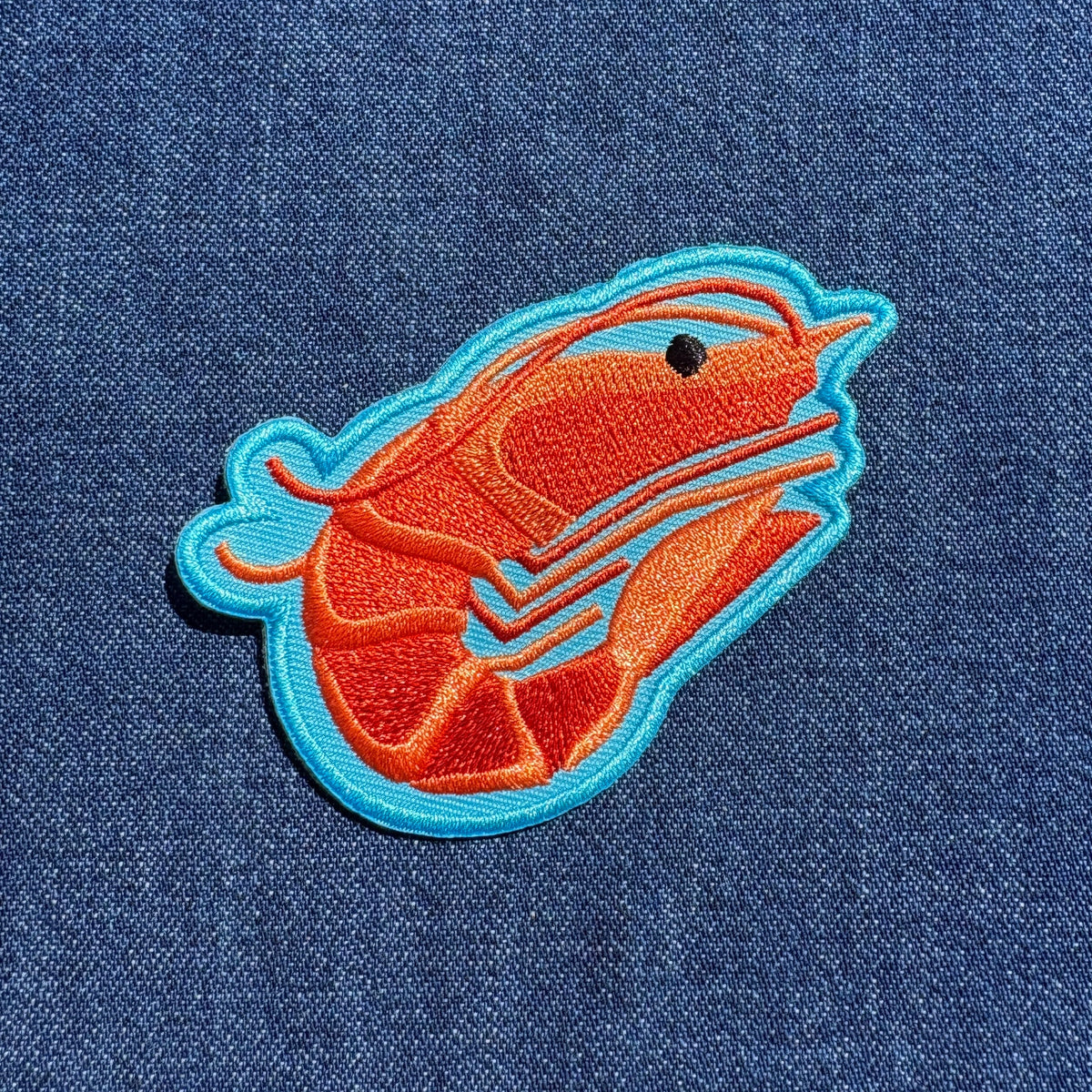 An embroidered shrimp patch in orange and blue on a blue denim fabric background.