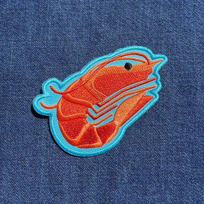 An embroidered shrimp patch in orange and blue on a blue denim fabric background.