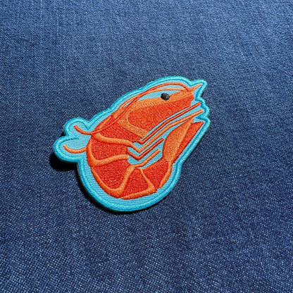 An embroidered patch of a stylized shrimp in orange and blue on a blue denim fabric background.