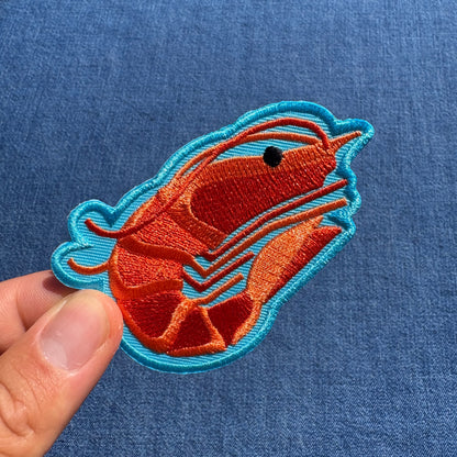 A close-up of a hand holding an embroidered shrimp patch against a blue denim fabric background.