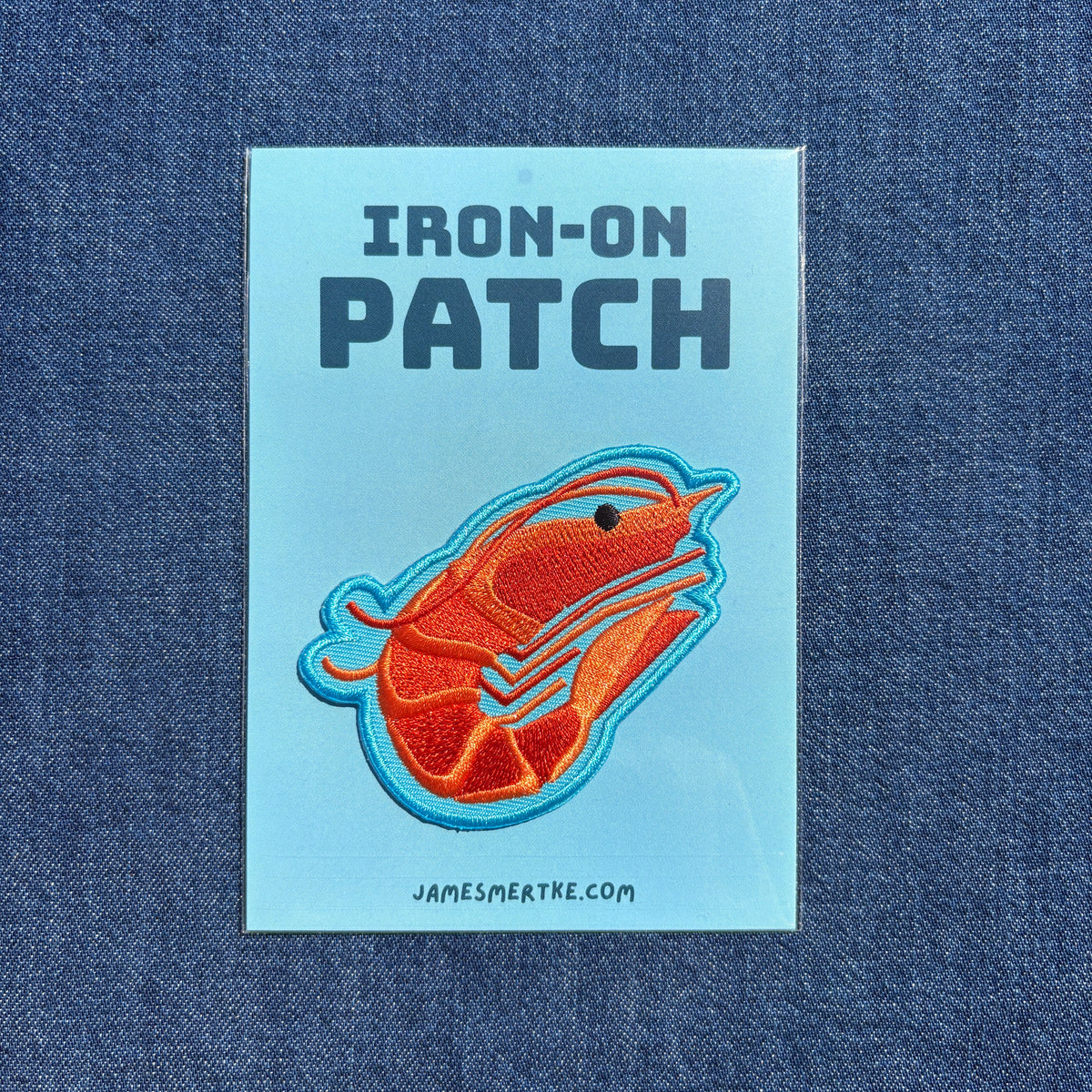 An iron-on patch featuring a stylized orange shrimp with a blue outline on a light blue backing card, displayed flat against a blue denim fabric background. The backing card reads 'IRON-ON PATCH' and 'JAMESMERTKE.COM'
