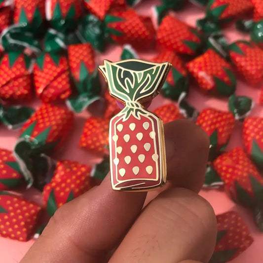 An enamel pin depicting a strawberry bon bon candy is half in front of a pink background with the real candies spread over. The enamel pin has shiny golden outlines with red and green coloring. 