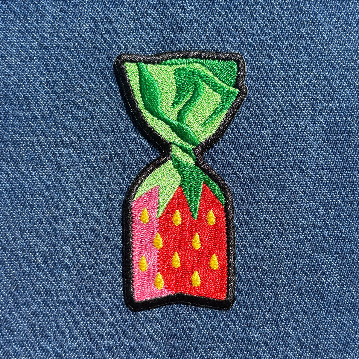 An embroidered patch shaped like a strawberry candy, featuring green leaves on top, a red body with yellow seeds, and a black stitched border, shown on a blue denim background.