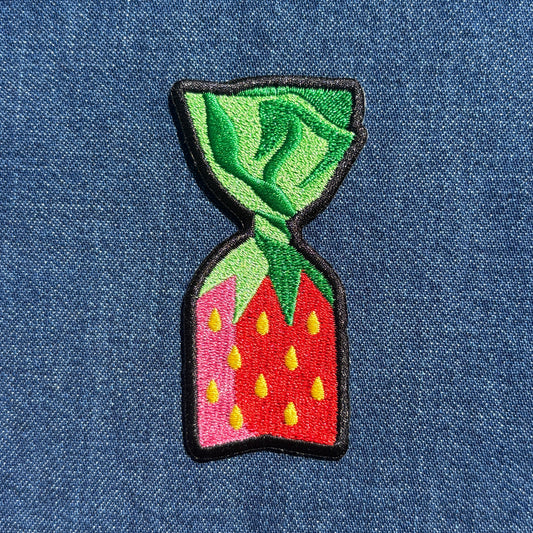 An embroidered patch shaped like a strawberry candy, featuring green leaves on top, a red body with yellow seeds, and a black stitched border, shown on a blue denim background.