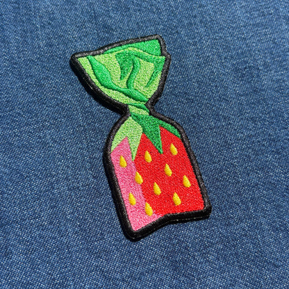 A colorful embroidered patch shaped like a strawberry candy, featuring green leaves, a red body with yellow seeds, and a pink side accent, all outlined in black and placed on a blue denim background.