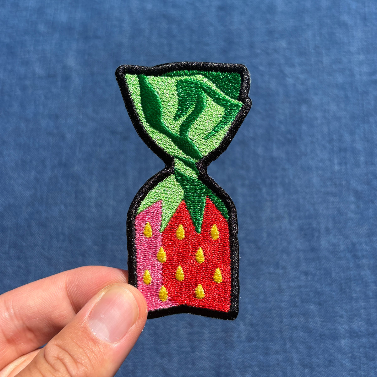 A hand holding an embroidered patch shaped like a strawberry candy, featuring green leaves, a red body with yellow seeds, outlined in black, against a blue textured background.