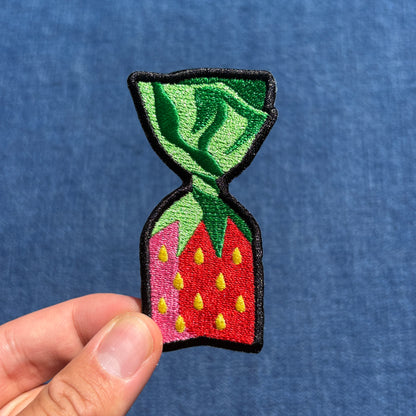 A hand holding an embroidered patch shaped like a strawberry candy, featuring green leaves, a red body with yellow seeds, outlined in black, against a blue textured background.