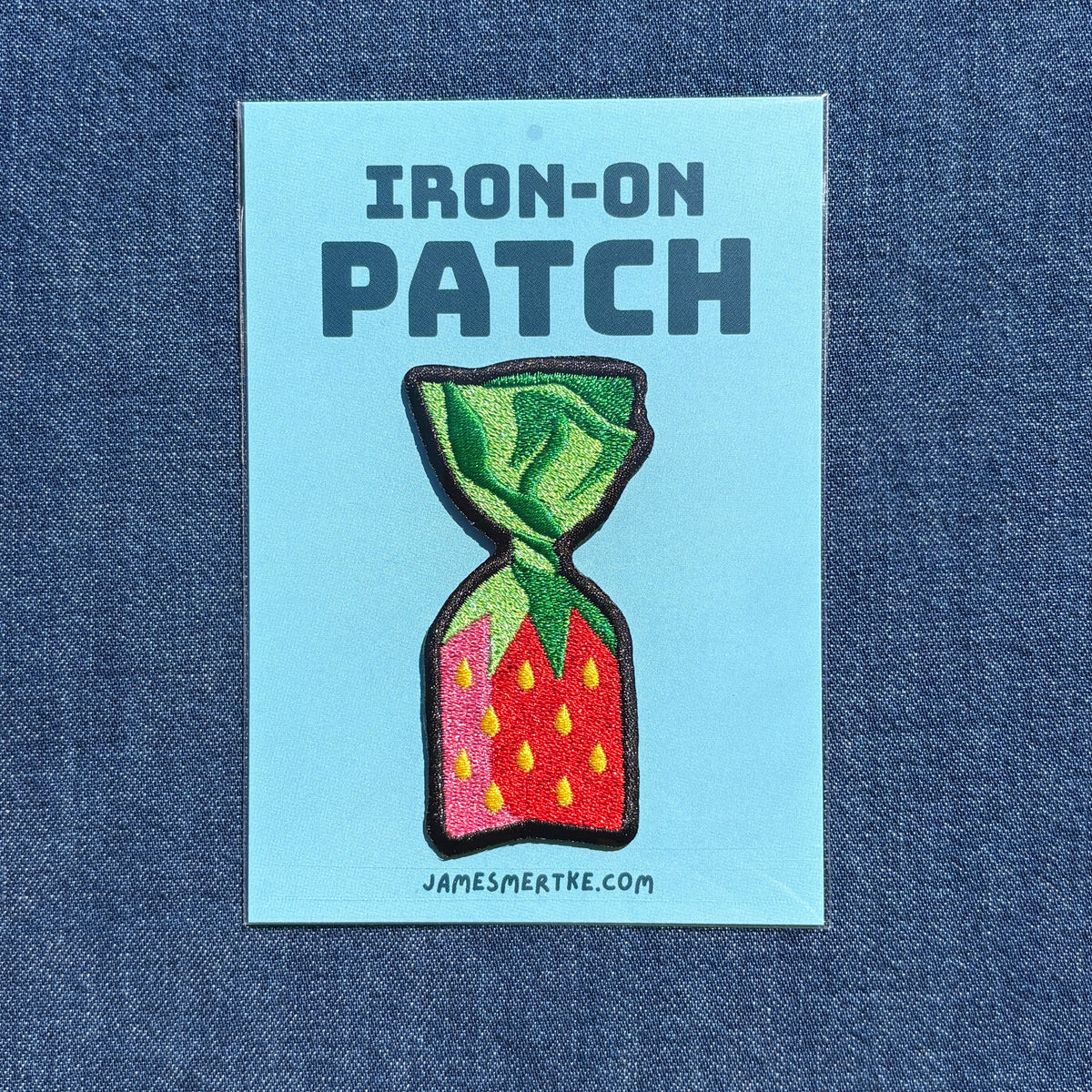 A rectangular light blue iron-on patch backing card on a blue denim background, featuring the text 'IRON-ON PATCH' above an embroidered strawberry candy design with green leaves, red/pink body, and yellow seeds. The card also shows 'JAMESMERTKE.COM'.