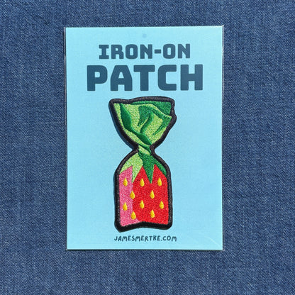 A rectangular light blue iron-on patch backing card on a blue denim background, featuring the text 'IRON-ON PATCH' above an embroidered strawberry candy design with green leaves, red/pink body, and yellow seeds. The card also shows 'JAMESMERTKE.COM'.