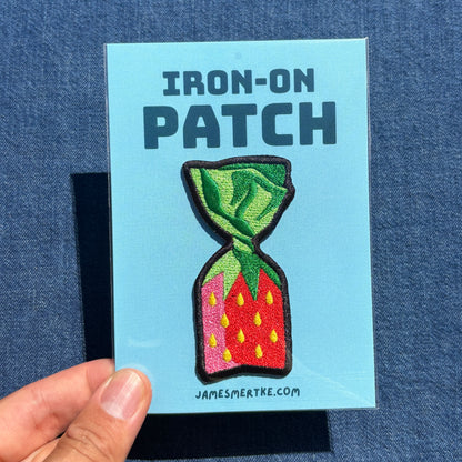 A hand holds a light blue card labeled 'IRON-ON PATCH' featuring an embroidered strawberry candy patch. The card also shows the website JAMESMERTKE.COM