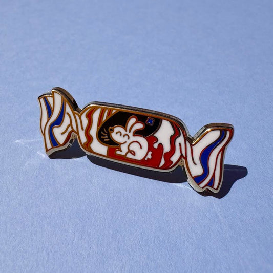 This enamel pin depicts a white rabbit candy with its red, white, and blue wrapper with a cheerful bunny. The pin is displayed on a flat blue background.