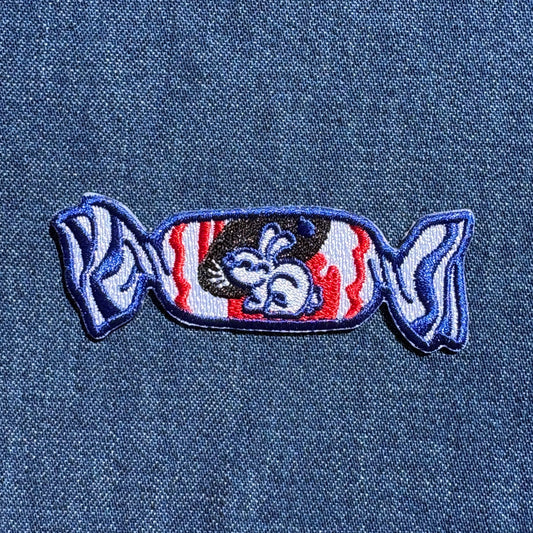 An embroidered candy-shaped patch featuring a stylized rabbit with red, white, and blue details, placed on a blue denim fabric background.