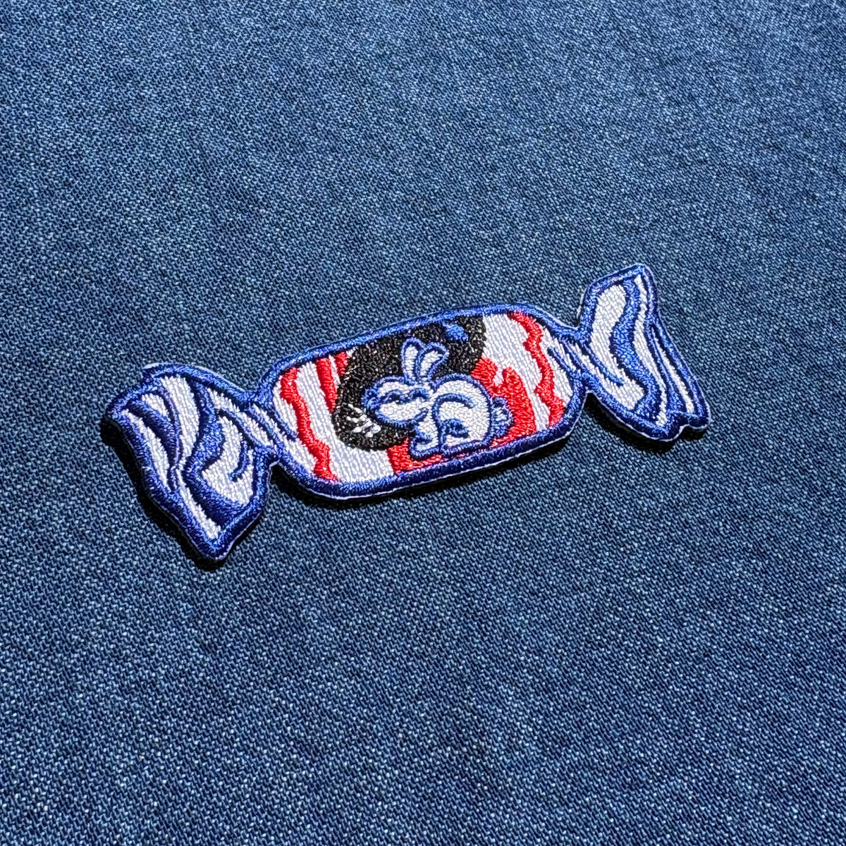 A candy-shaped embroidered patch with a blue border, red and white striped wrapper, and a black center featuring a white bunny, shown on a blue denim fabric background.