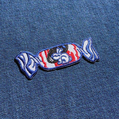 A candy-shaped embroidered patch with a blue border, red and white striped wrapper, and a black center featuring a white bunny, shown on a blue denim fabric background.