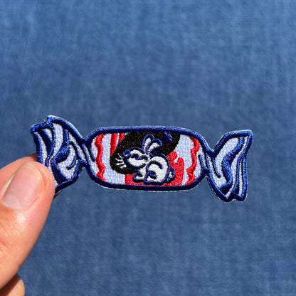 A small embroidered patch shaped like a candy, held by a person's fingers against a blue fabric background. The patch features a cartoon rabbit with red and white wavy stripes behind it and blue outlines.