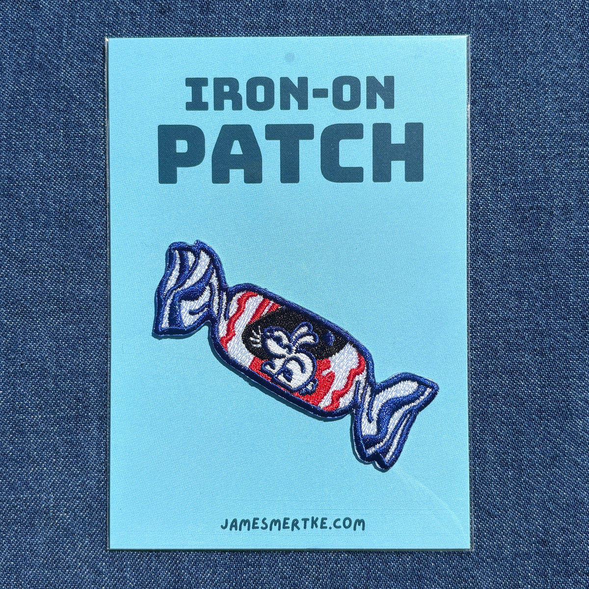An iron-on white rabbit candy patch on a light blue backing card, displayed on a dark blue denim fabric background. The backing card features the text 'IRON-ON PATCH' and the website 'JAMESMERTKE.COM'.