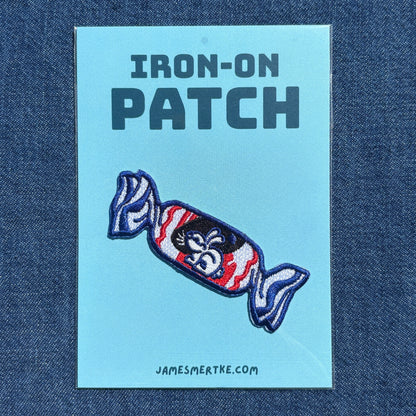 An iron-on white rabbit candy patch on a light blue backing card, displayed on a dark blue denim fabric background. The backing card features the text 'IRON-ON PATCH' and the website 'JAMESMERTKE.COM'.