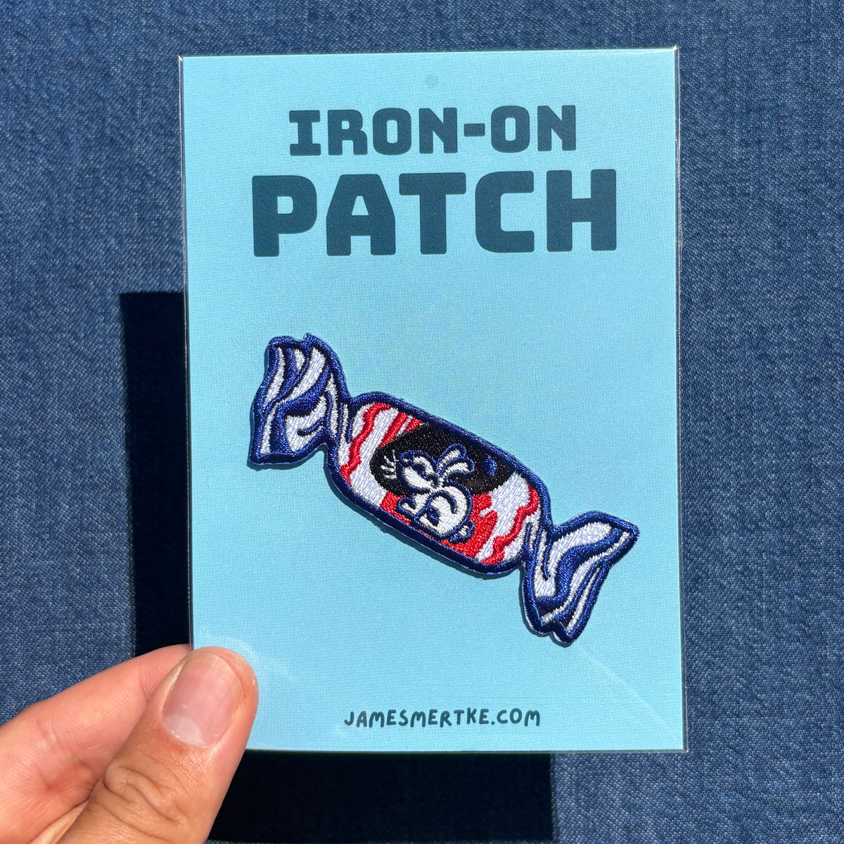 A hand holds a light blue backing card that reads 'IRON-ON PATCH' above an embroidered candy-shaped patch with red, white, black, and blue white rabbit candy. The card is photographed against a dark blue textured fabric background.